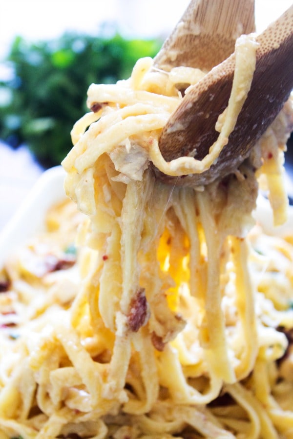 Chicken Fettuccine Bake – Filled to the brim with Alfredo sauce, tossed with chicken, bacon & sun-dried tomatoes, ready in just 45 minutes. photo of two wooden spoons grabbing Chicken Fettuccine Bake from dish