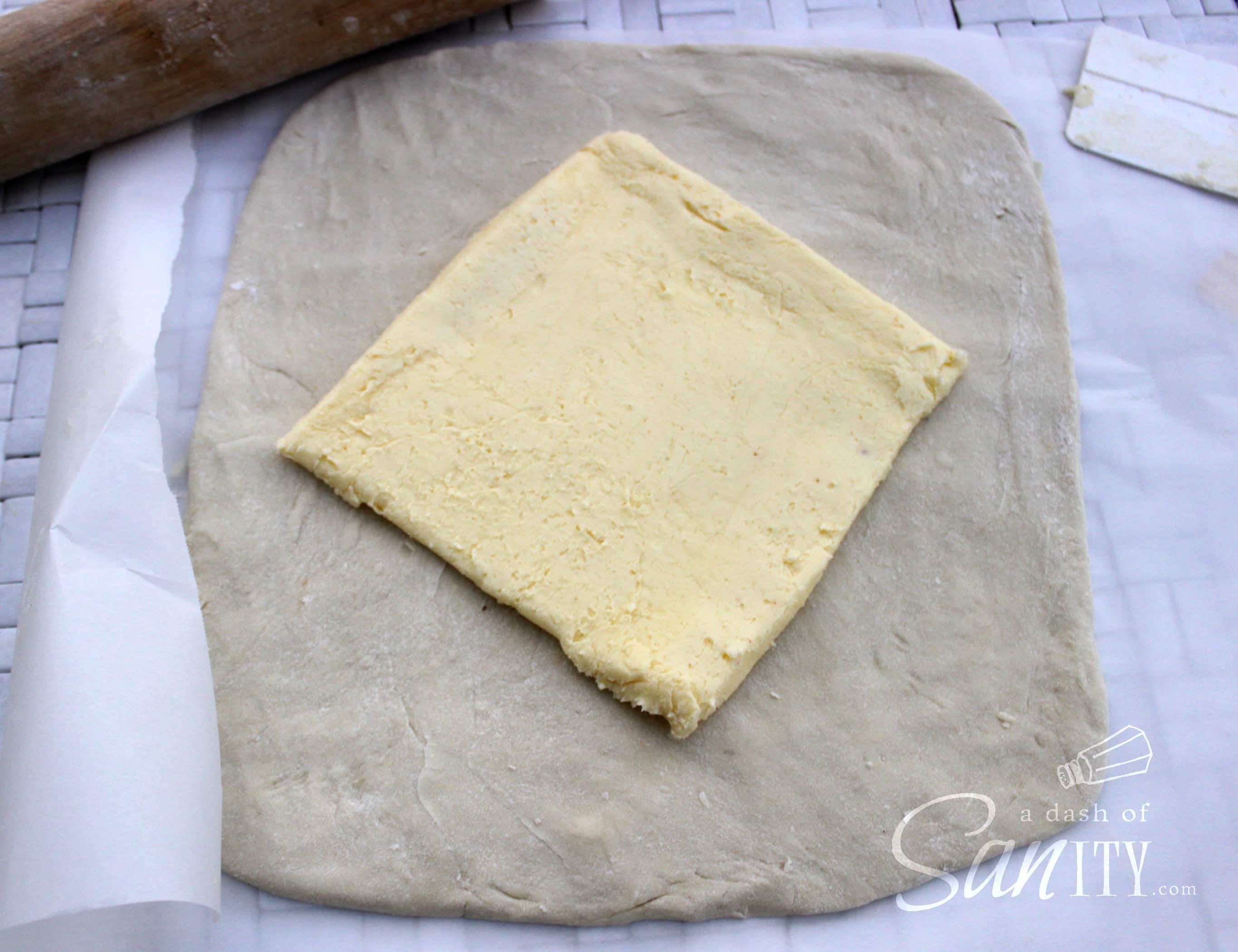 These Chocolate Croissants are light, flaky, buttery croissants with a deliciously rich dark chocolate center. A perfect treat for any chocolate lover. croissant dough rolled into a rectangle with a square sheet of butter on top, on parchment paper with a wooden rolling pin