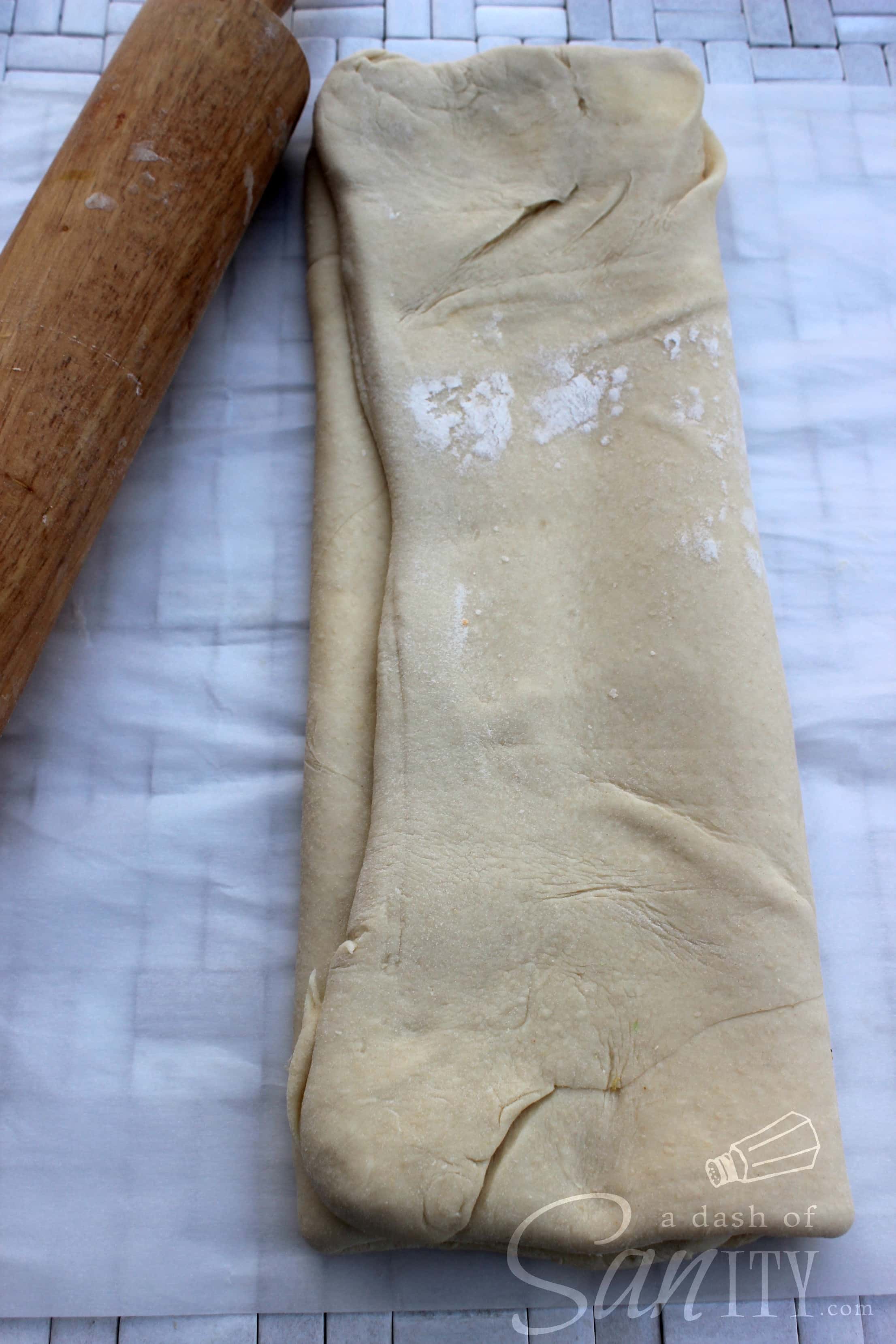 These Chocolate Croissants are light, flaky, buttery croissants with a deliciously rich dark chocolate center. A perfect treat for any chocolate lover. croissant dough rolled out into rectangle and then folded into thirds, on parchment next to a rolling pin