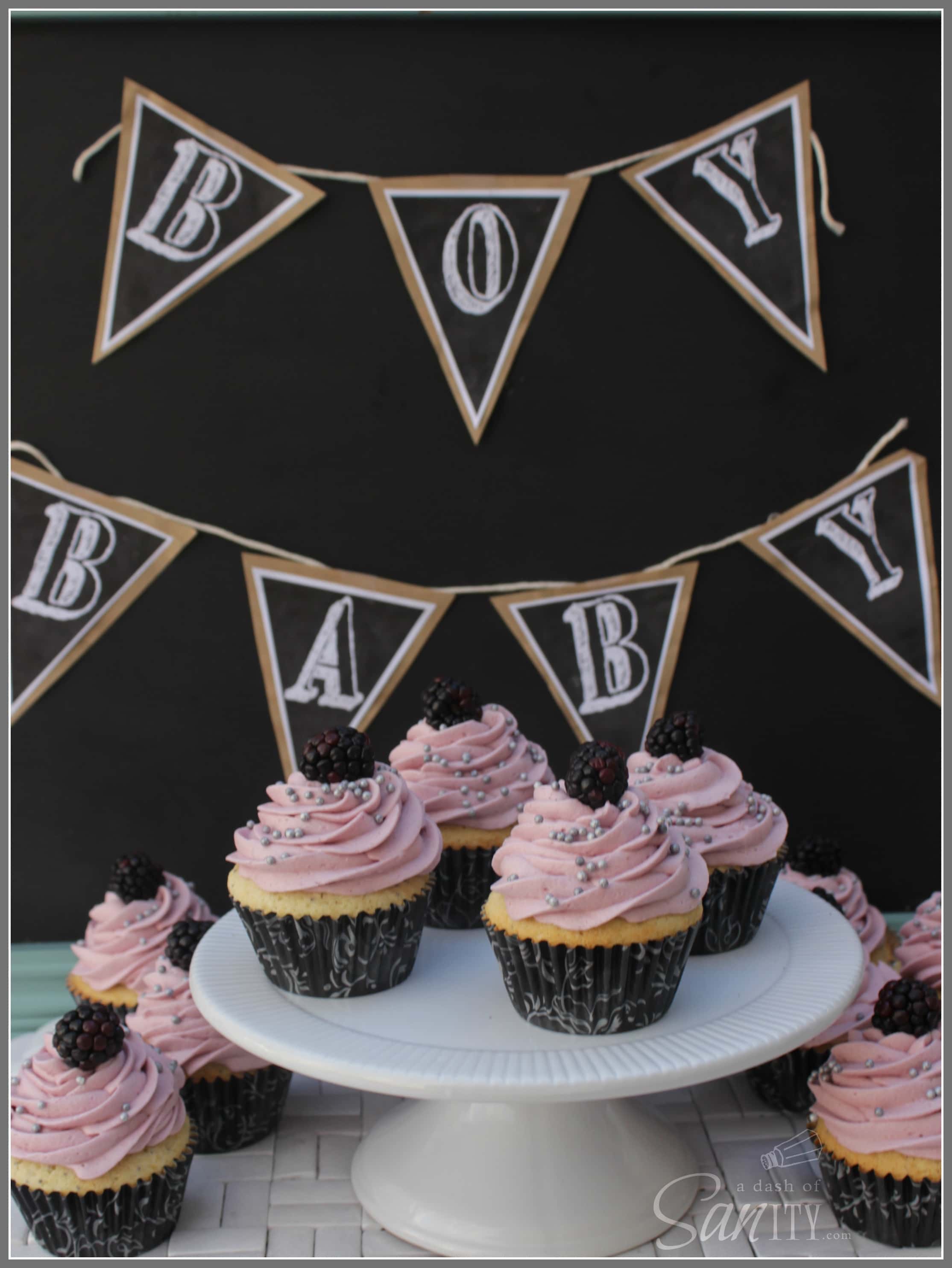 These Lemon-Poppy Seed Cupcake with Blackberry White Chocolate Buttercream are not only sweet to the eye but also to the palate. 'Lemon-Poppy Seed Cupcake with Blackberry White Chocolate Buttercream on a cake stand and counter, with a banner above that reads "BABY BOY"