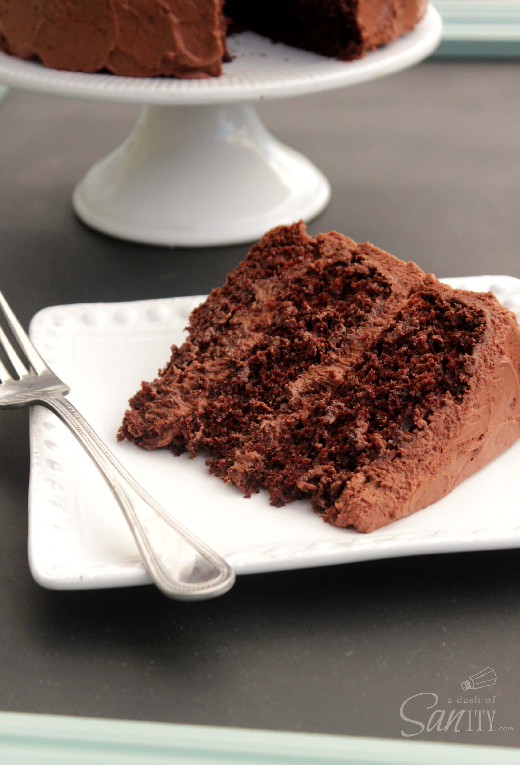 Best Ever Chocolate Layer Cake slice on a square plate, with cake on cake stand in the background