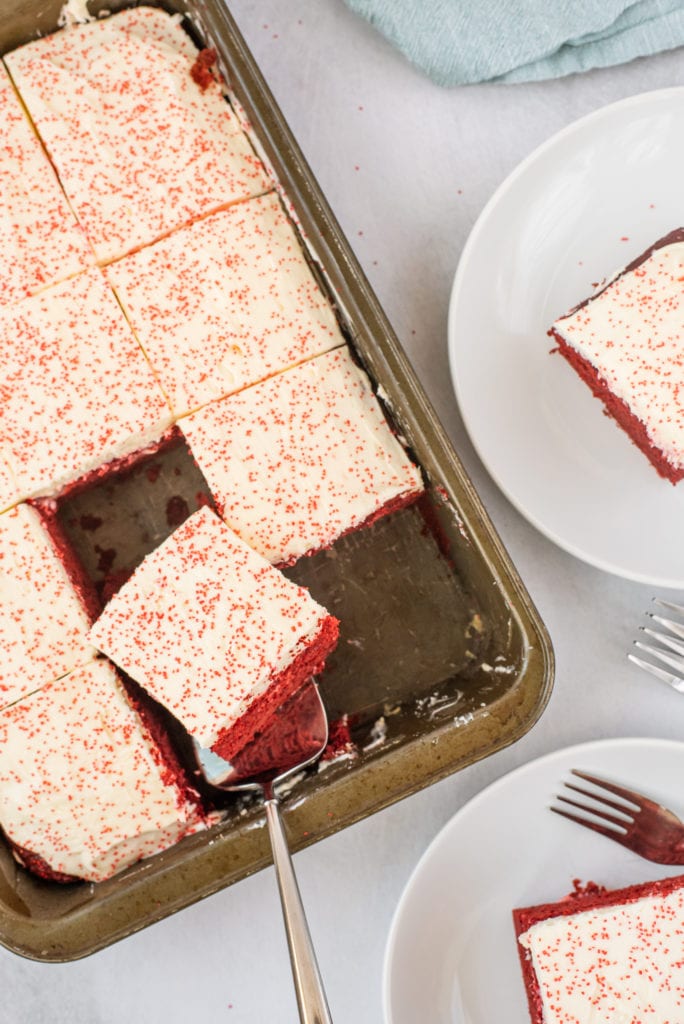 overhead photo of a red velvet cake in a pan with slices being removes