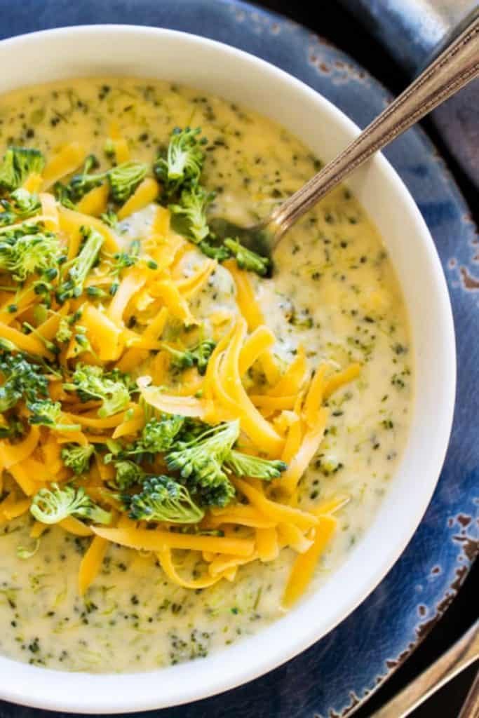 Broccoli Cheese Soup in a white bowl with a spoon, garnished with broccoli and shredded cheese