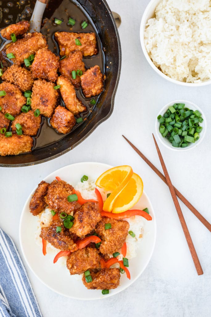overhead photo of a plate of orange chicken with a skillet and bowl of rice