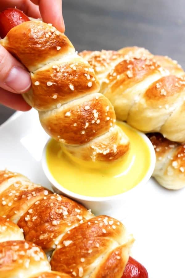 Simple & delicious these EASY PRETZEL HOT DOGS are the perfect combination a hot dog and a soft pretzel- a kid favorite in our home. photo of pretzel hot dogs on a plate, dipping one into a small bowl of honey mustard sauce