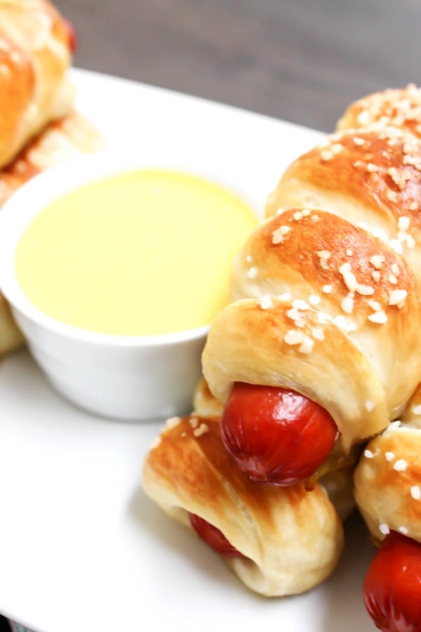 Simple & delicious these EASY PRETZEL HOT DOGS are the perfect combination a hot dog and a soft pretzel- a kid favorite in our home. photo of pretzel hot dogs stacked on a white plate with side of honey mustard dipping sauce