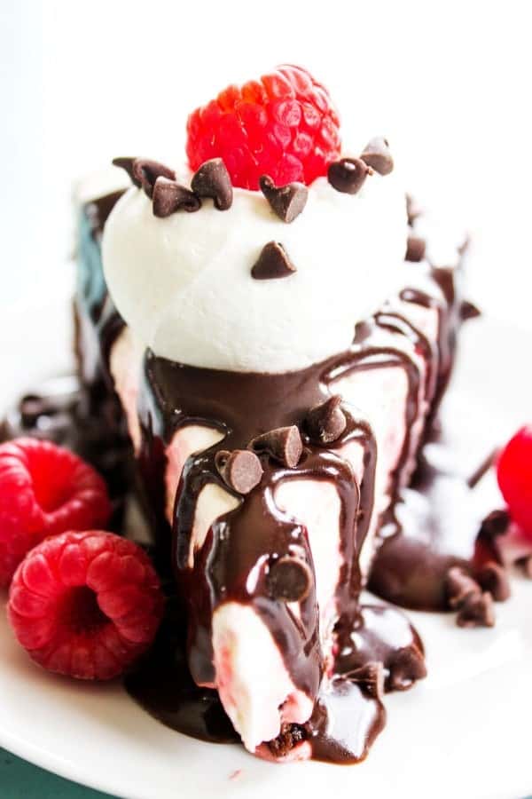 FUDGE BROWNIE NO BAKE RASPBERRY CHEESECAKE - the best ever hot fudge sauce drizzled over a fresh raspberry no bake cheesecake all on top of a fudgy delicious brownie. This is the dessert you have been craving. FUDGE BROWNIE NO BAKE RASPBERRY CHEESECAKE - White plate, blue table