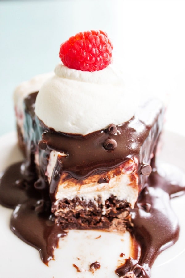 FUDGE BROWNIE NO BAKE RASPBERRY CHEESECAKE - the best ever hot fudge sauce drizzled over a fresh raspberry no bake cheesecake all on top of a fudgy delicious brownie. This is the dessert you have been craving. slice of brownie cheesecake on plate and bite out of it