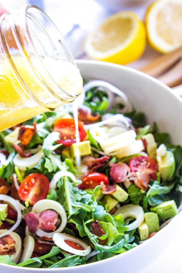 ARUGULA AND HEARTS OF PALM SALAD - this Brazilian inspired salad is rich with flavor and dressed in a light, sweet lemon vinaigrette. dressing in a mason jar being poured over arugula and hearts of palm salad in a white bowl