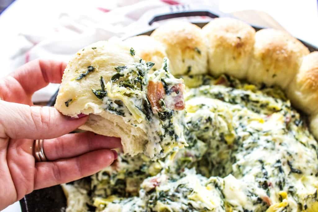 SPINACH ARTICHOKE BACON DIP RECIPE- creamy, delicious and easy to prepare this is a party must have. Adding bacon to your classic spinach artichoke dip is a fantastic twist that all your friends & family love. SPINACH ARTICHOKE BACON DIP RECIPE- Bite from dish displayed over iron skillet on a wooden cutting board