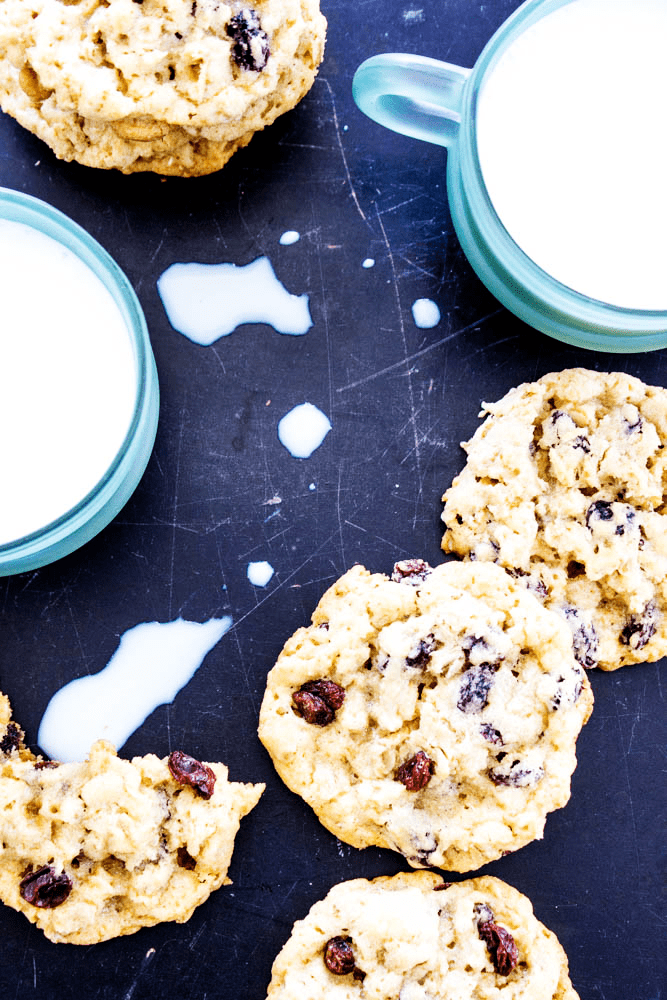Oatmeal Raisin Cookies laid out on counter with glass mugs of milk, one cookie with bites taken