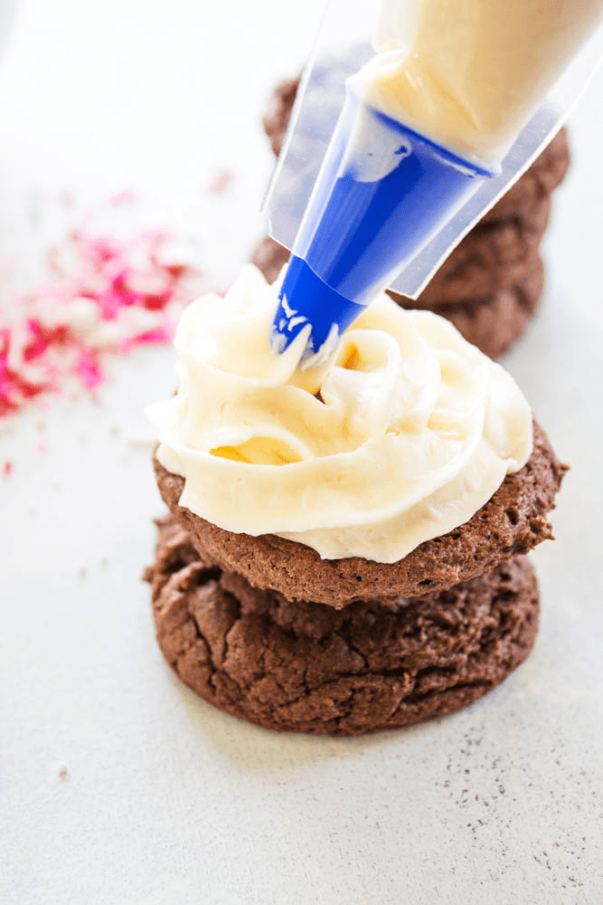 EASY WHOOPIE PIE RECIPE- Soft and chewy Devil's Food cake cookies sandwiched with cream cheese frosting, these whoopie pies are out of this world. piping bag piping cream cheese frosting onto a chocolate cake cookie