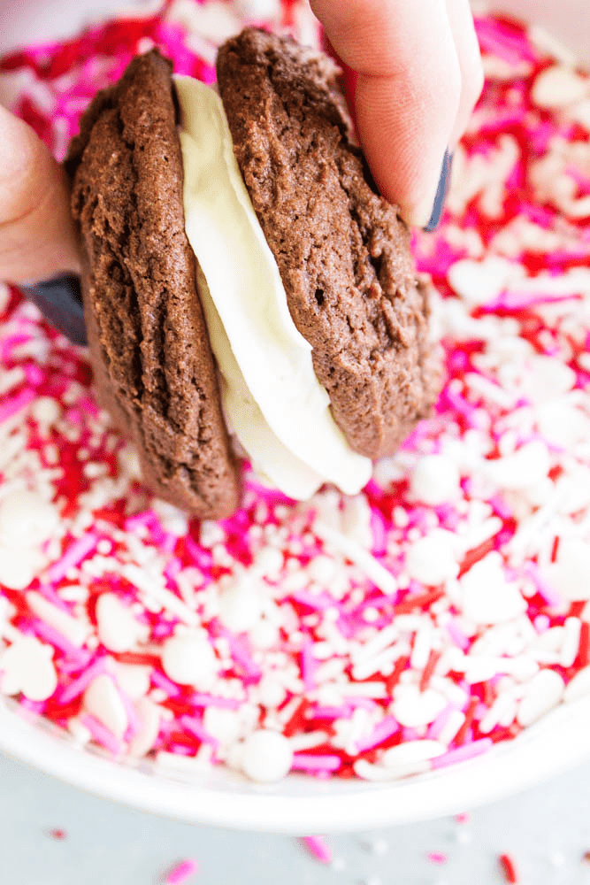 EASY WHOOPIE PIE RECIPE- Soft and chewy Devil's Food cake cookies sandwiched with cream cheese frosting, these whoopie pies are out of this world. rolling a whoopie pie cookie frosting in red, pink, and white sprinkles