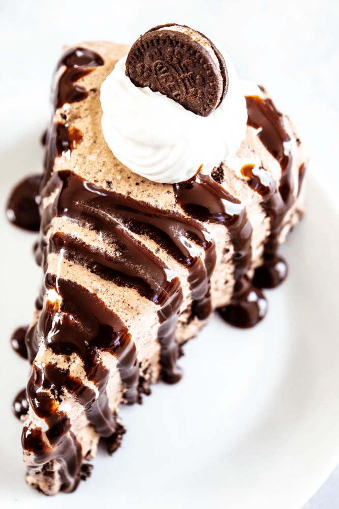 chocolate oreo ice cream cake slice on a plate served with hot fudge sauce, whipped cream, and a mini oreo