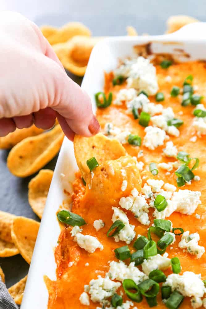 buffalo chicken dip topped with green onions and blue cheese in a rectangle dish, with a hand dipping a frito in 