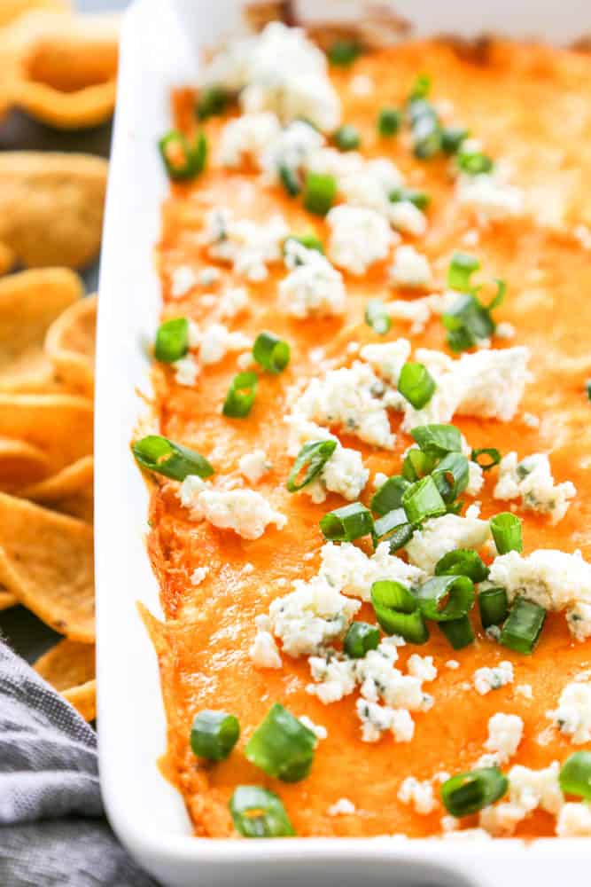 Buffalo Chicken Dip served in white baking dish topped with sliced green onions and blue cheese. with fritos on side