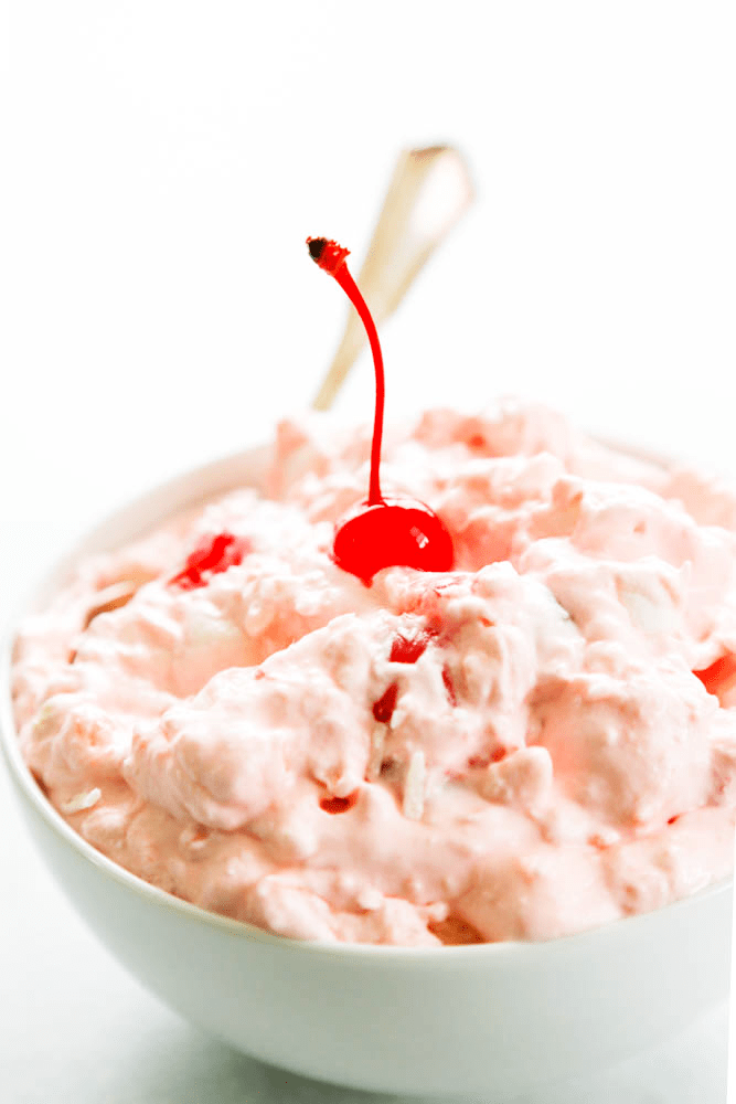 Pink Pineapple Fluff topped with a maraschino cherry in a bowl with a spoon