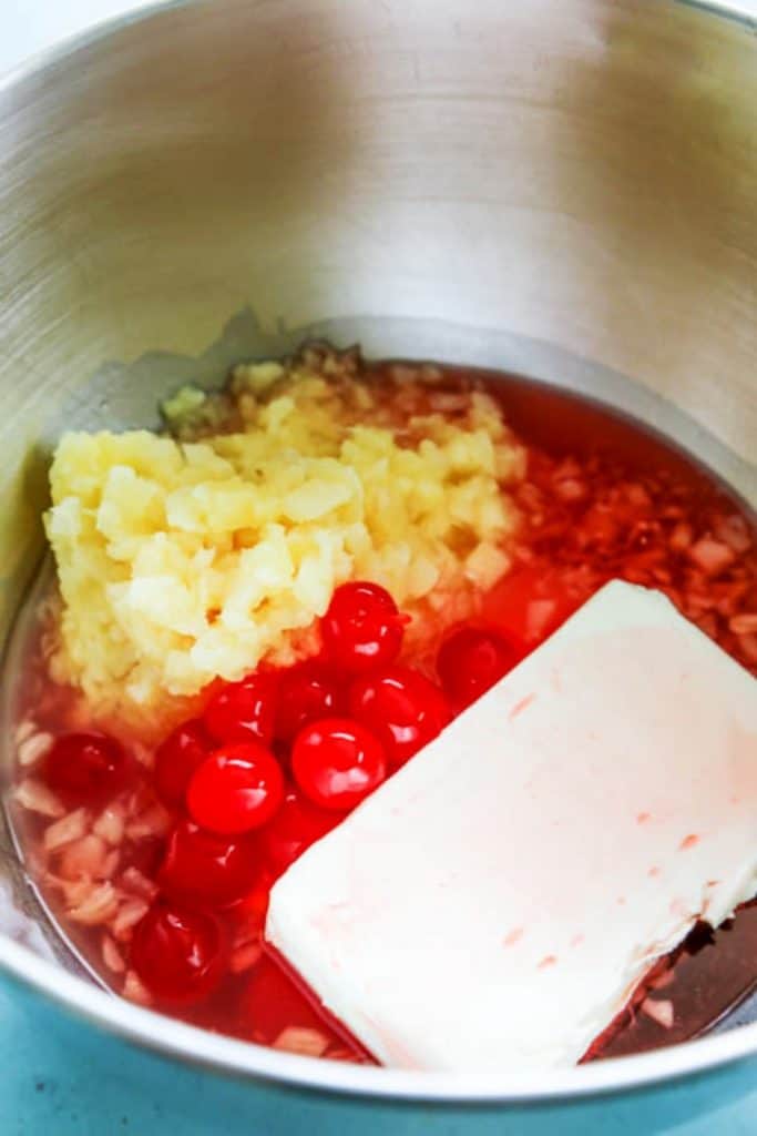 Pink Pineapple Fluff ingredients in a metal mixing bowl, pineapple, cherries, and cream cheese block