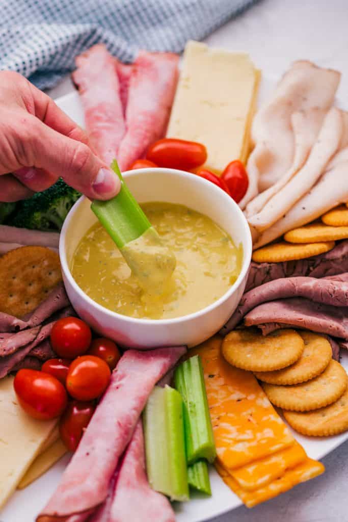 carolina mustard dip in a white bowl in center of a plate filled with meats, cheeses, crackers, and veggies with celery being dipped in sauce