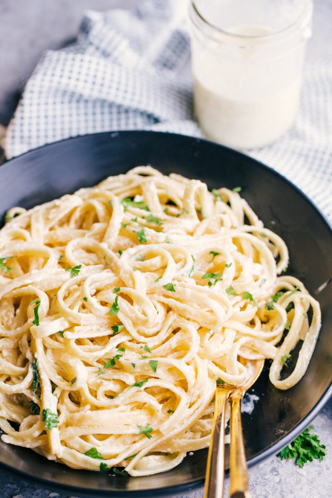 This easy FETTUCCINE ALFREDO recipe is simply delicious. Cheesy, creamy and made from scratch, this sauce is the perfect comfort food made at home. Fettuccine Alfredo served in a black bowl with glass of alfredo sauce on table