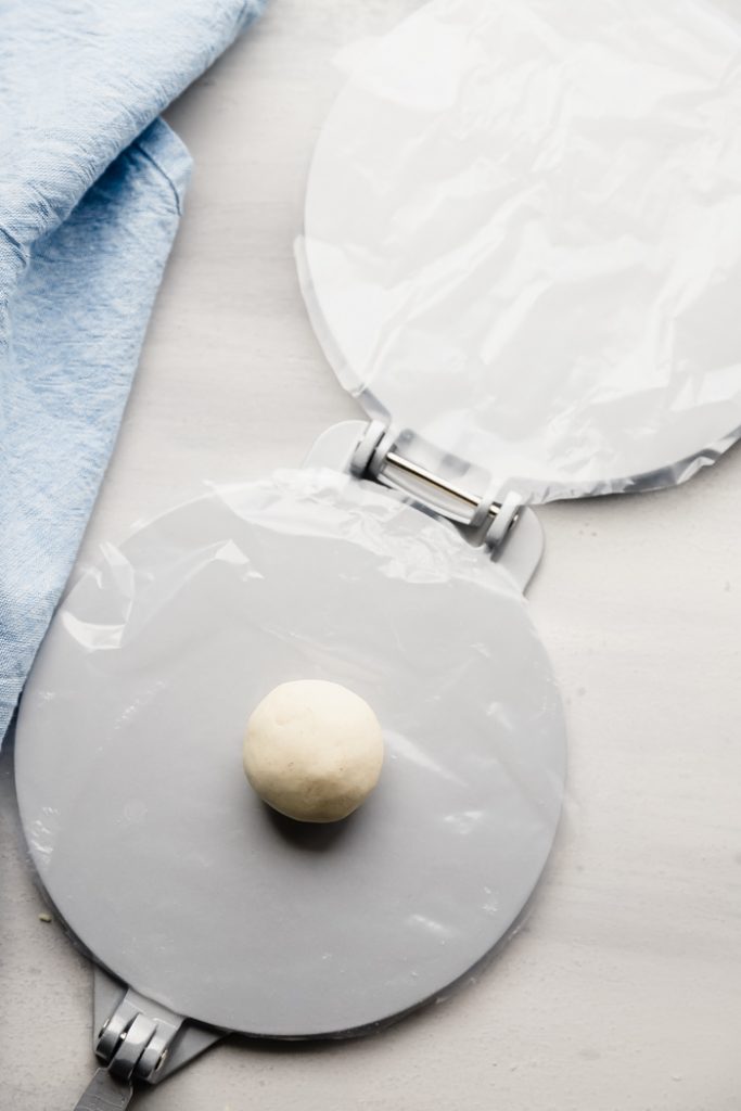 photo of an open tortilla press with both sides lined with plastic wrap, with a ball of dough in the middle