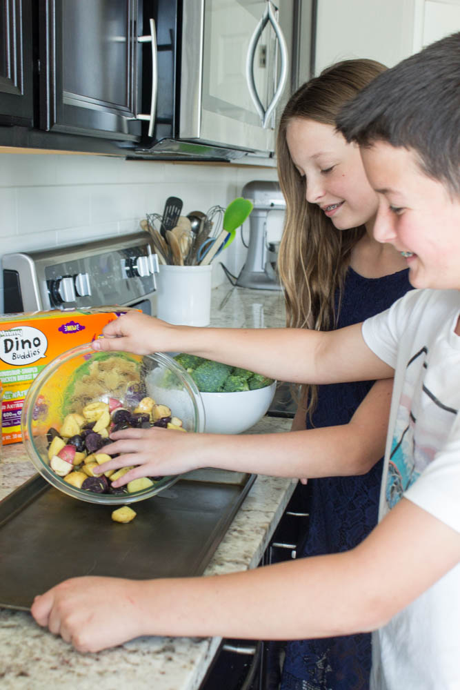 photo of 2 kids pouring halved and seasoned mini potatoes from a glass bowl onto sheet pan 