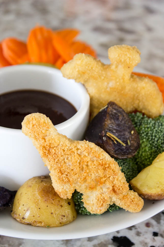 photo with cut potatoes, dino nuggets, cut carrots and broccoli with a small dipping bowl of barbecue sauce on a plate