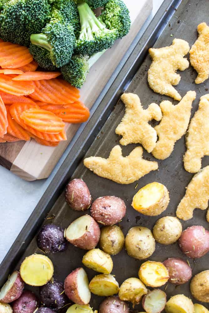 overhead photo of cut mini potatoes and dino nuggets on a sheet pan next to a cutting board with wavy cut carrots and broccoli florets