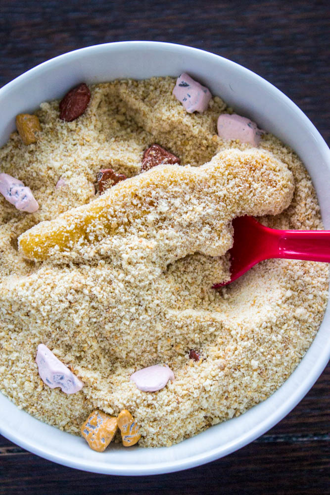 photo showing t-rex dino nugget buried in breadcrumb "sand" in a white bowl, with candy rocks and a red spoon for digging