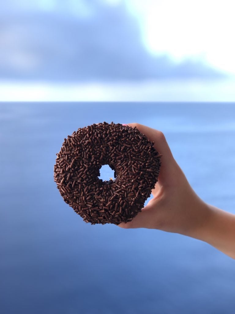 photo of a chocolate donut with chocolate sprinkles being held up, with ocean in background