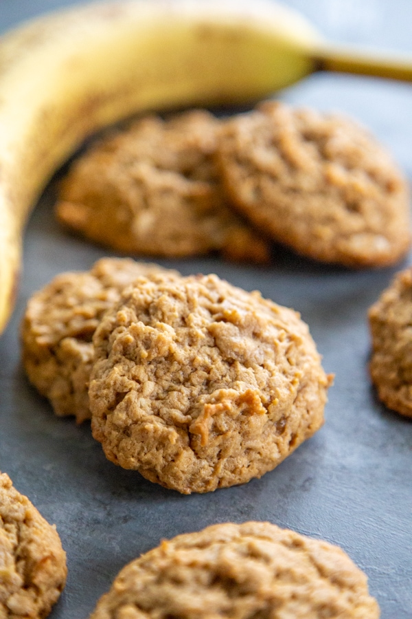 Chewy HEALTHY PEANUT BUTTER BANANA COOKIES are a twist on your peanut butter cookie recipe. Made with no butter and less sugar, they make a great snack! cookies on a work surface with a banana in the background.