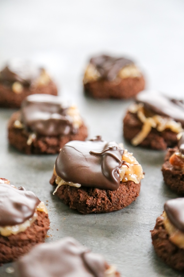 These Mini German Chocolate Thumbprint Cookies are fudgey-brownie like cookies topped with a chocolate drizzle, toasted coconut, and chopped pecans. Thumbprint cookies lined up on a baking sheet.