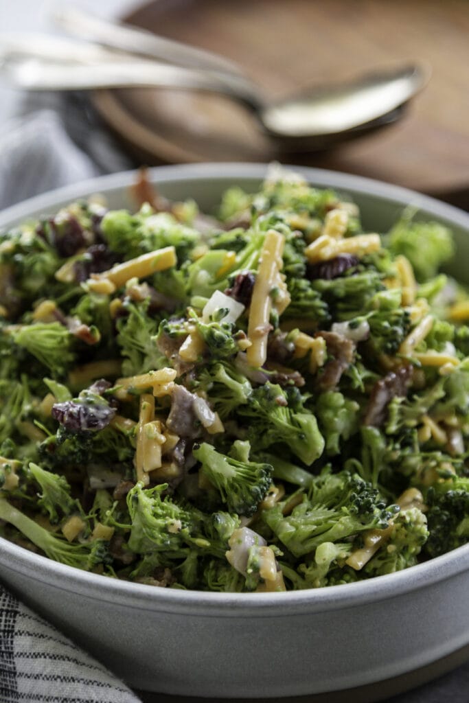 bowl of fresh broccoli salad with spoons and wooden plate in back drop